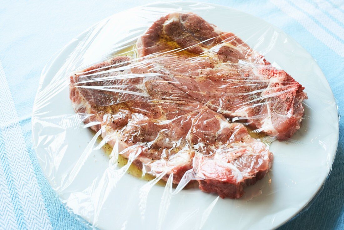 Zwei Schweinenackensteaks in Olivenölmarinade auf weißem Teller unter Frischhaltefolie