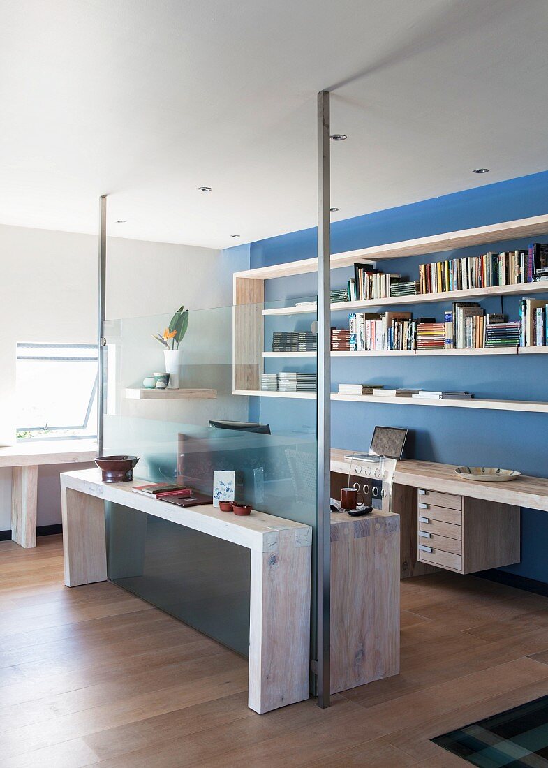 Half-height glass wall with steel supports between living area and office space with shelving on blue-painted wall