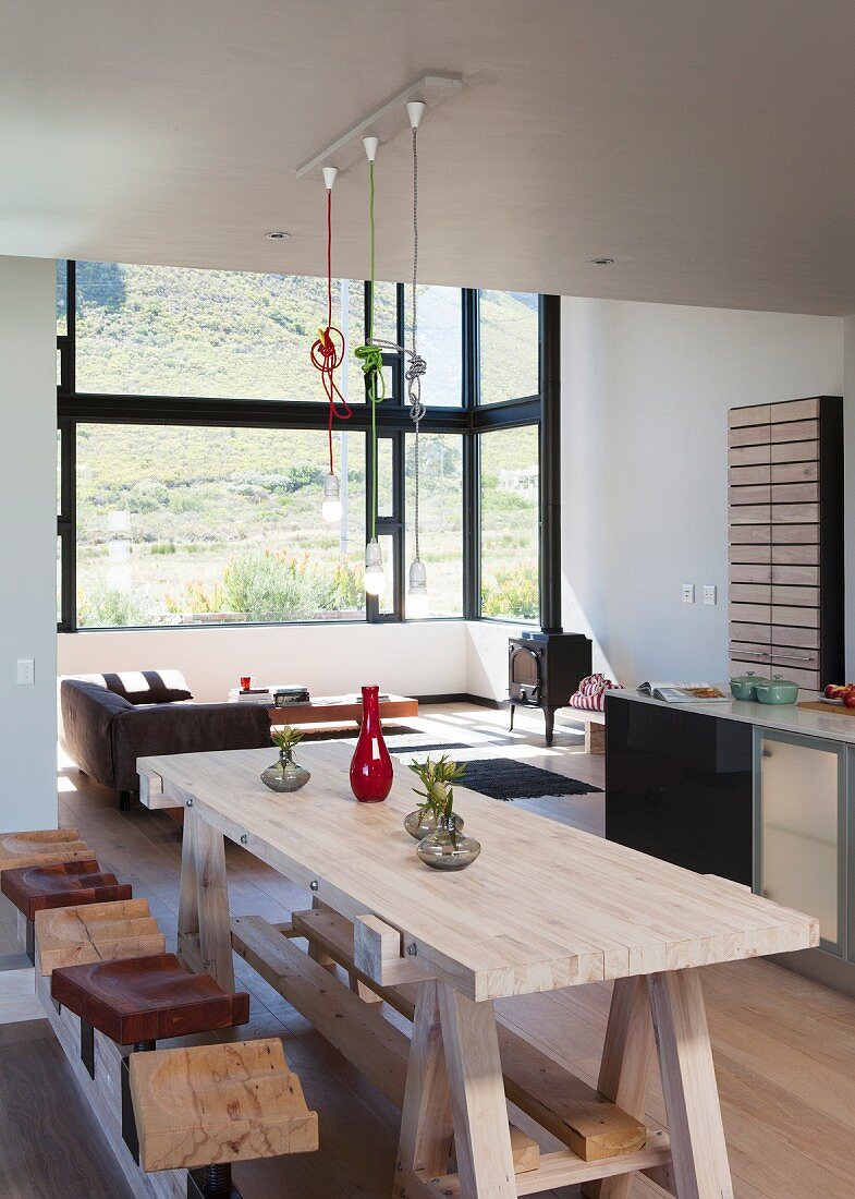 Wooden, workbench-style dining table and stools in open-plan interior; lounge area in panoramic bay window