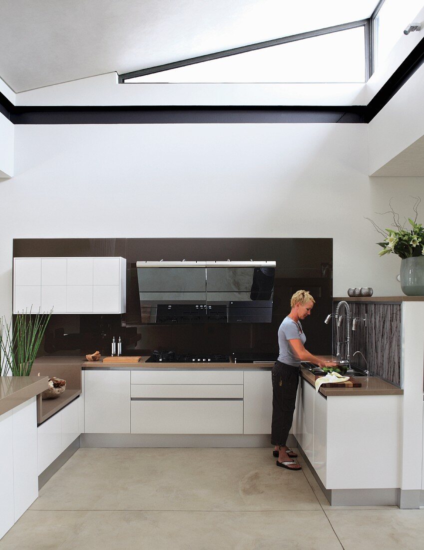 Open-plan, white, designer fitted kitchen with triangular transom light below ceiling; woman at kitchen counter