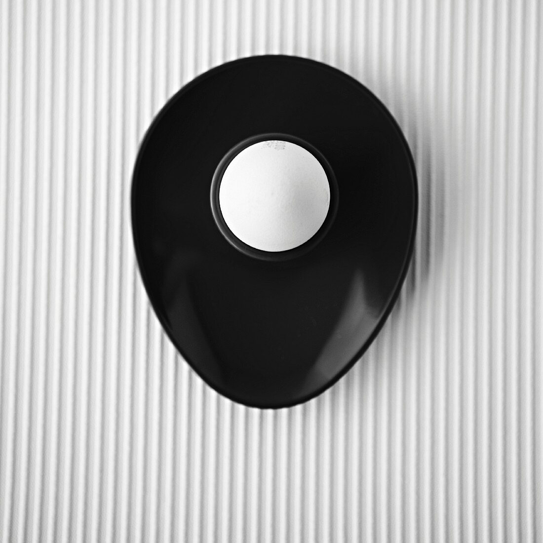 A white egg in a black egg cup, overhead, on a white textured background