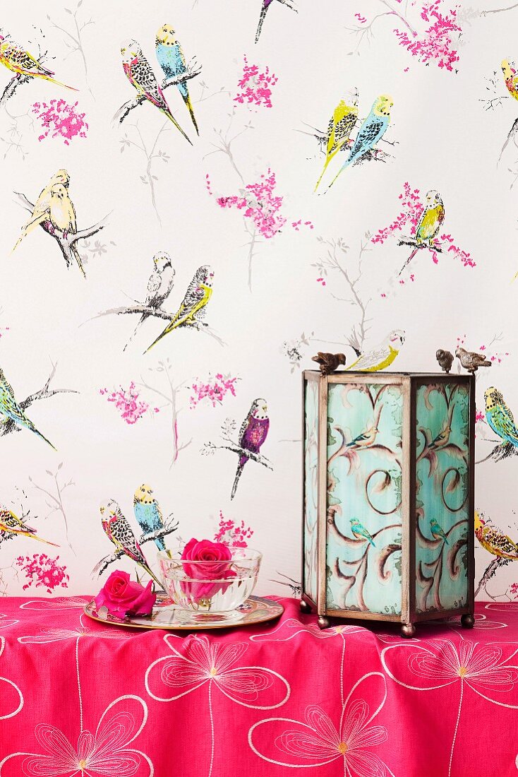 Decorative candle lantern on pink cloth against wall with whimsical, budgerigar-patterned wallpaper