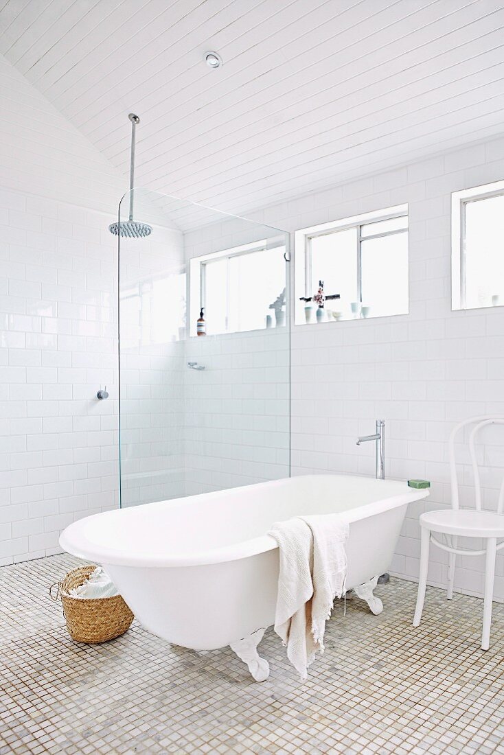 Free-standing bathtub and white-painted Thonet chair next to floor-level rainfall shower with glass partition