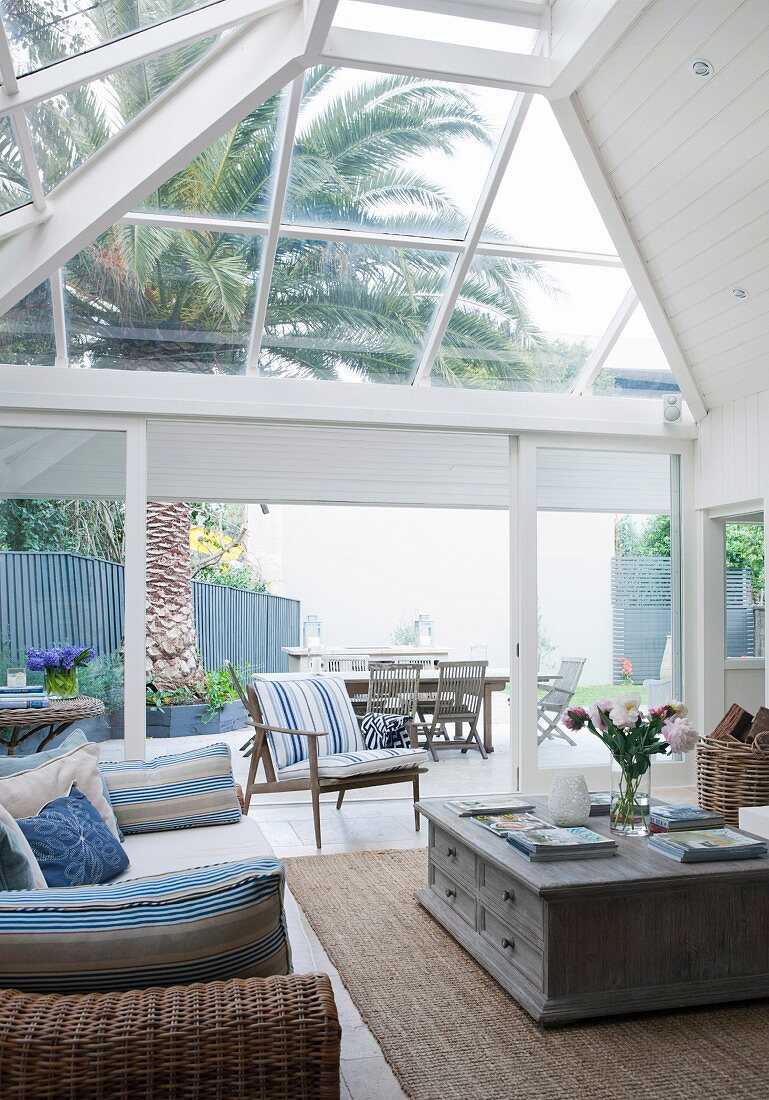 Wicker sofa with striped cushions in conservatory extension made from white-painted wood; view of terrace with large palm tree and wooden furniture