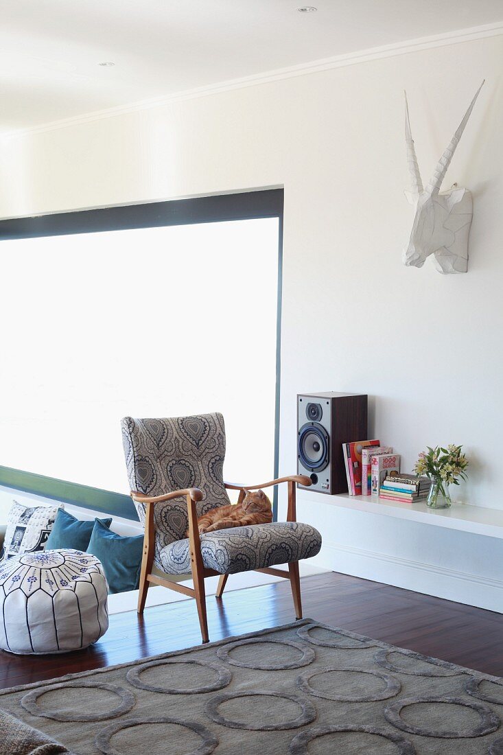 Pouffe and retro wooden armchair next to speaker and books on shelves; hunting trophy sculpture on wall