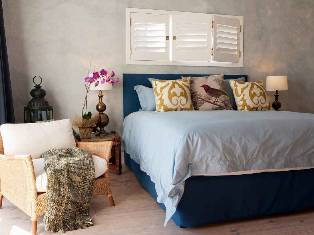 Double bed with frame and headboard upholstered in blue below window with closed interior shutters and rattan armchair
