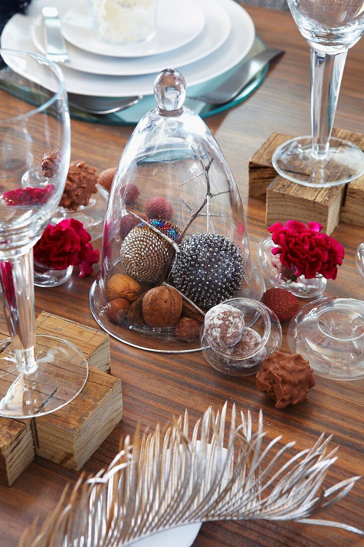 Nuts and decorative baubles under a glass cloche and pralines, flowers and a feather on a table decorated for Christmas