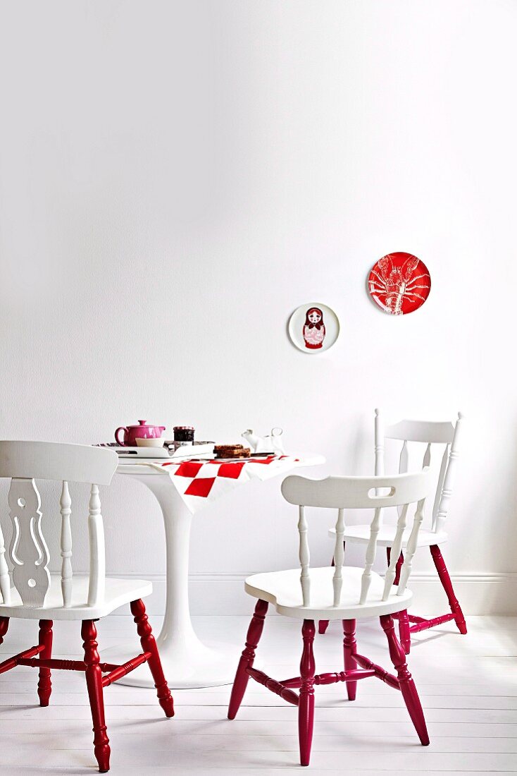 Alte Holzstühle weiß-rot gestrichen auf weißem Dielenboden vor weißer Wand, in der Mitte ein runder gedeckter weißer Tisch