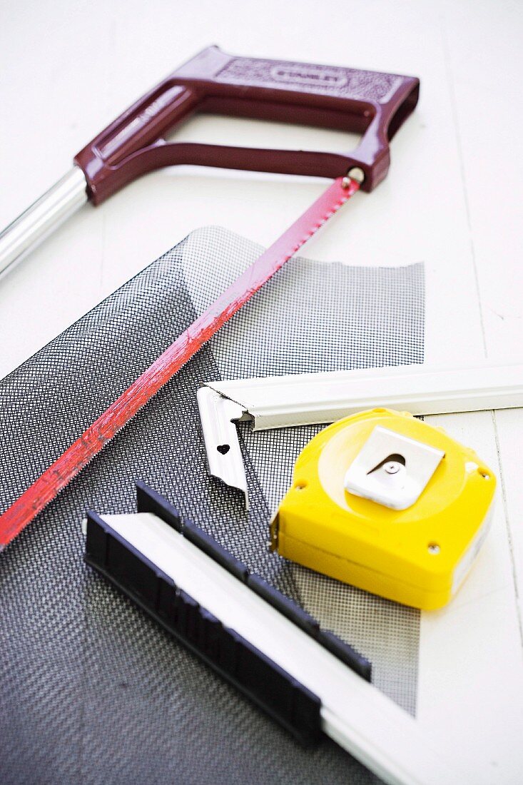 Red hacksaw, grey mesh, yellow tape measure and pieces of metal frame on white worksurface