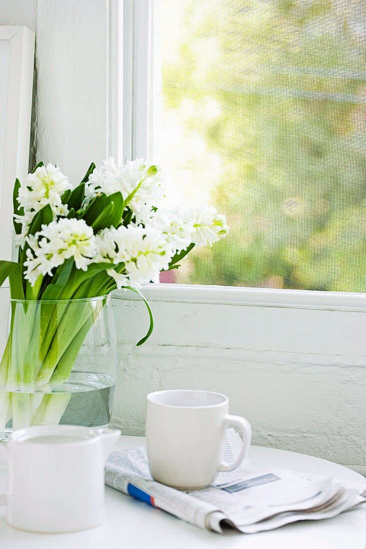 Frühstücksambiente eines Singles mit Kaffeetasse auf gefalteter Zeitung, daneben ein weißer Hyazinthenstrauß in einer Glasvase vor Vintagefenster