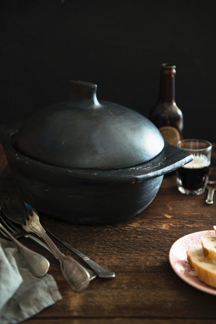 A black cooking pot, silver cutlery, bread and beer on a wooden table (England)