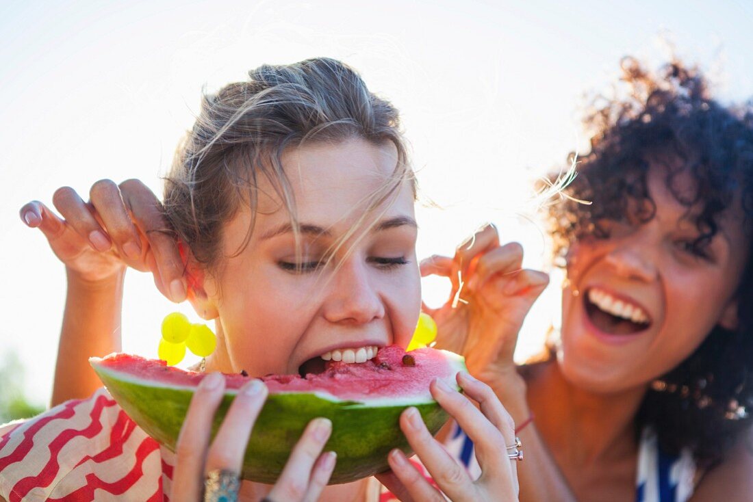A woman eating a wedge of melon while her friend holds grapes up by her ears as earrings