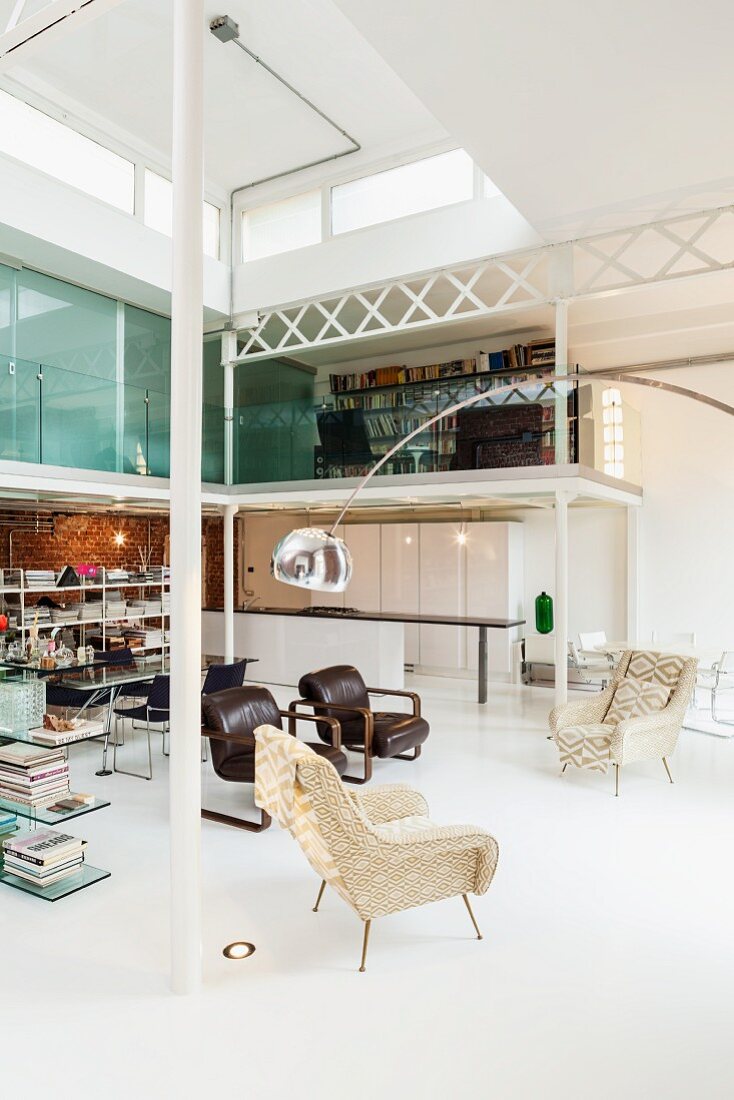 Spacious loft apartment with partition shelving, group of armchairs with arc lamp and modern kitchen below sleeping area on gallery; natural light falling through lantern skylight