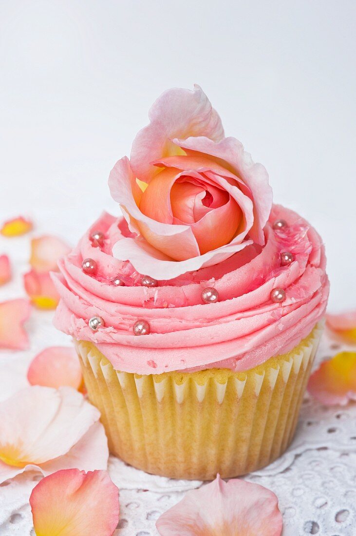 A pink cupcake decorated with rose petals and silver balls