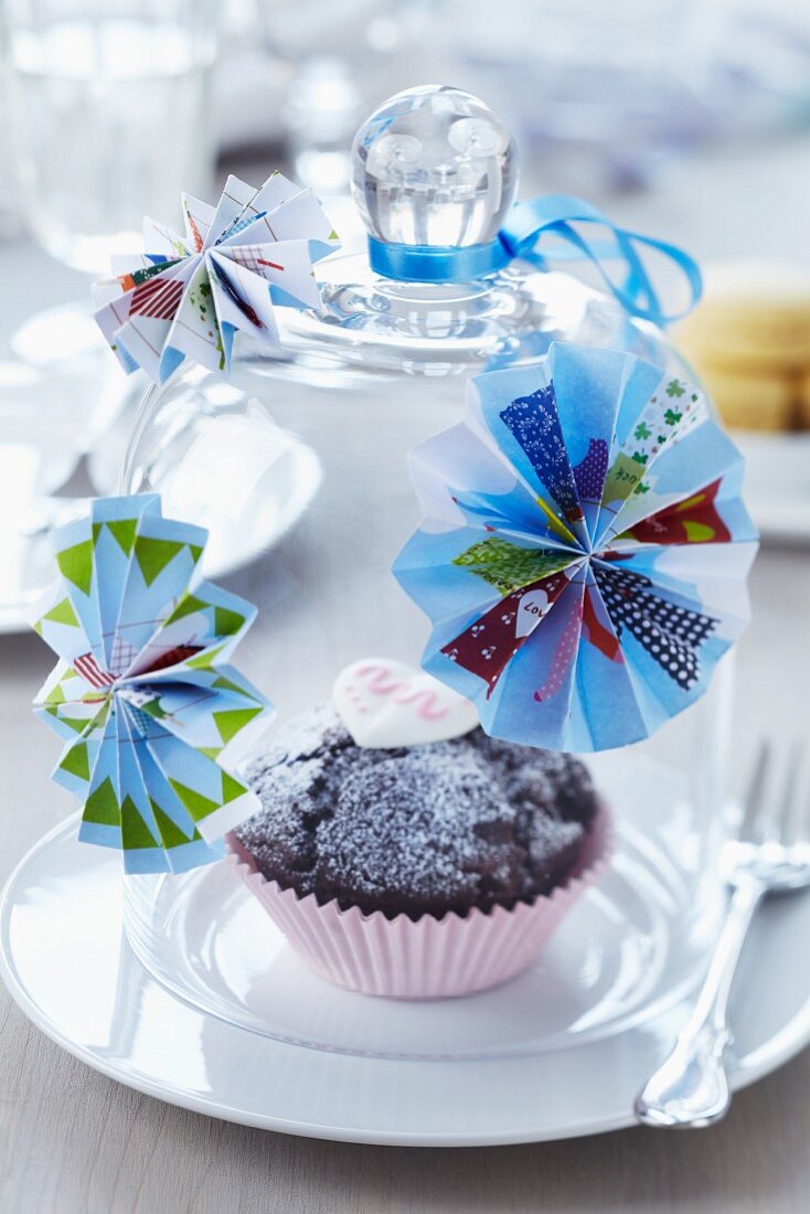 Muffin in pink paper case below glass cover decorated with paper rosettes