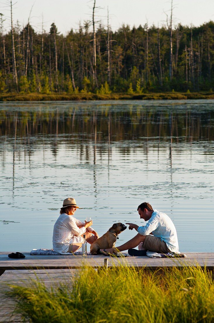 Man, woman and dog sitting on wooden jetty next to lake in evening sun