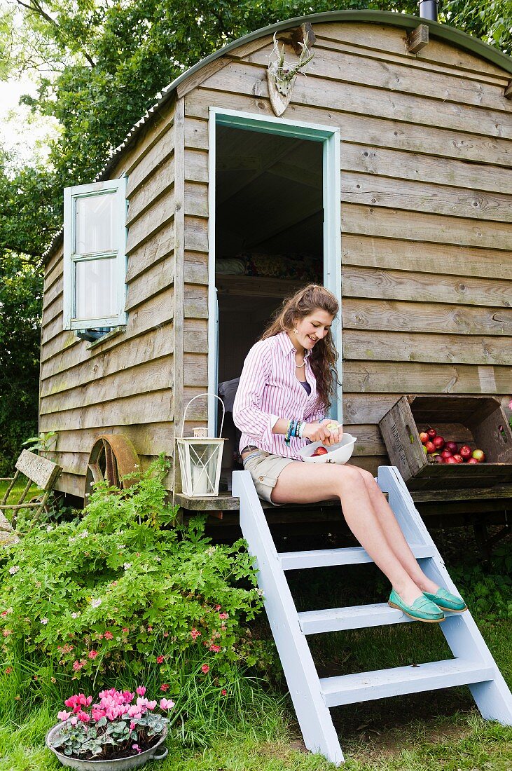 Woman sitting next to box of apples on pale blue ladder leading to nostalgic shepherds' hut amongst greenery