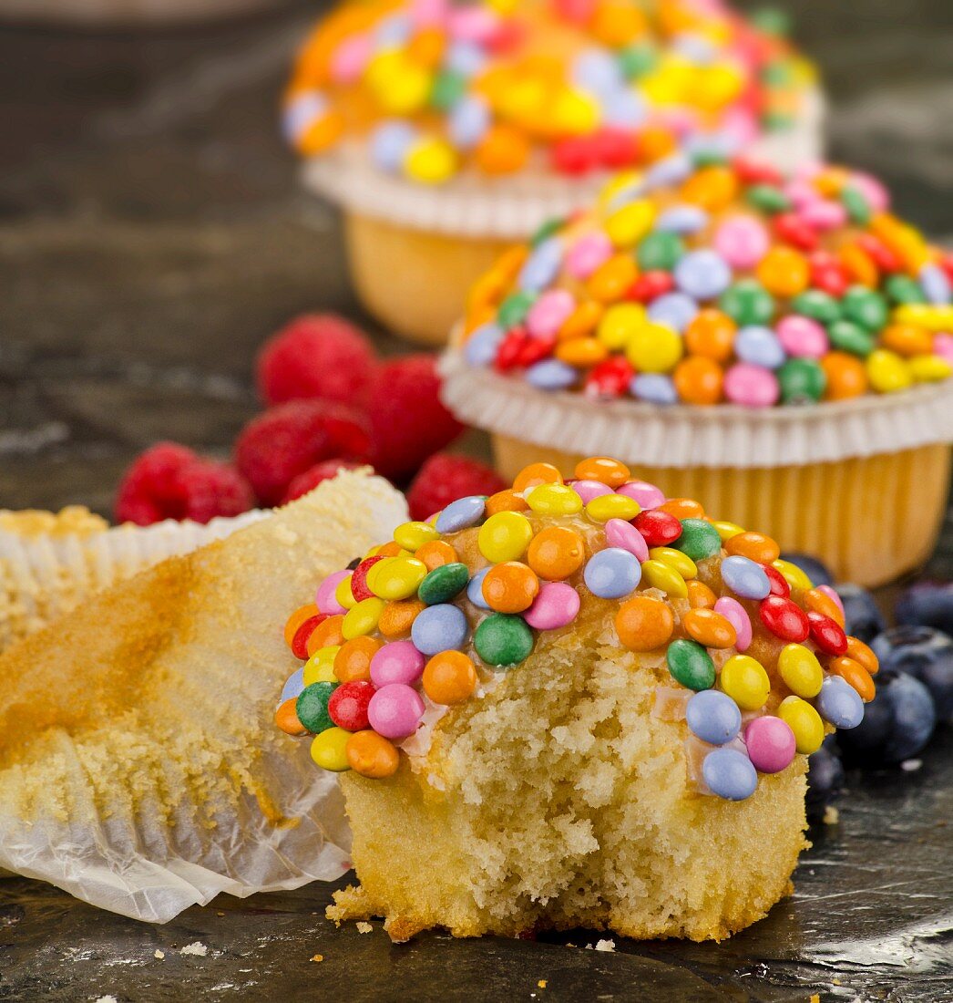Cupcakes decorated with colourful chocolate beans, whole and with a bite missing