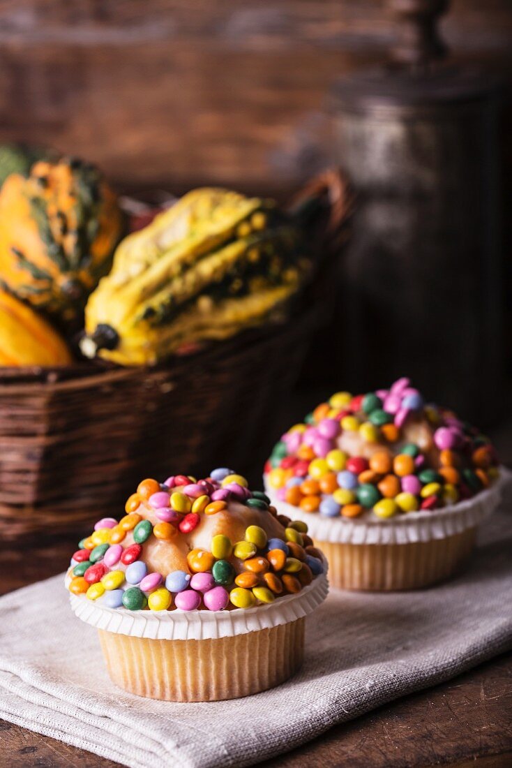 Cupcakes with colourful chocolate beans in front of a basket of squash