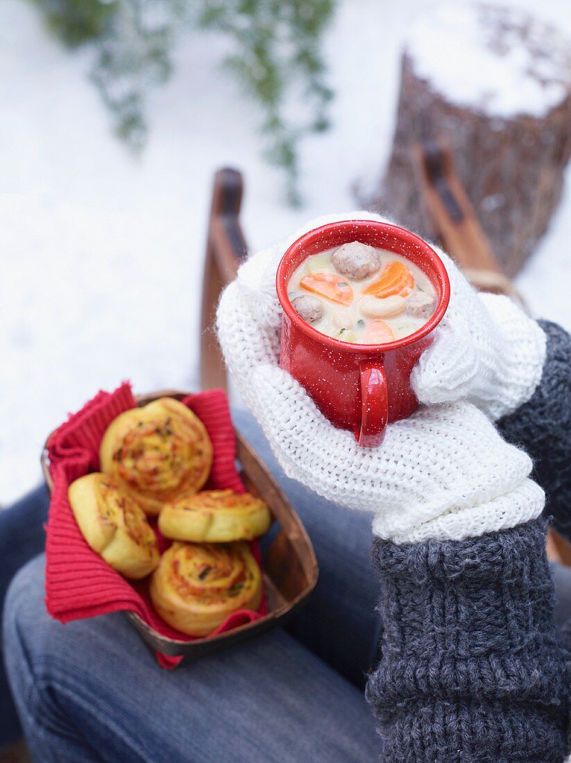Hands holding an enamel mug of bean and potato soup with meat dumplings with a basket of pumpkin and leek whirls