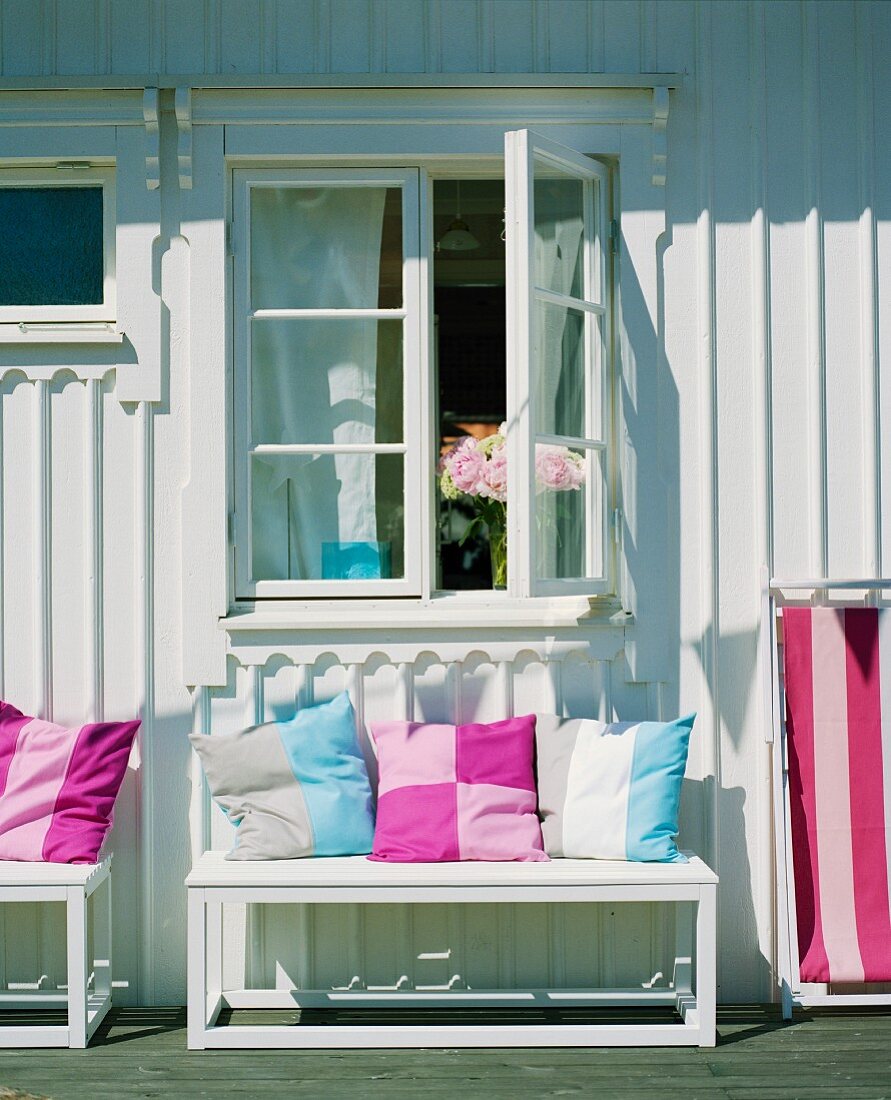 Colourful scatter cushions on bench outside white-painted wooden house on sunny day