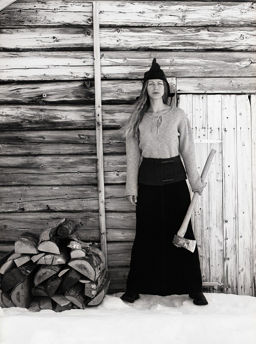 Woman with menacing stare holding axe standing against outside wall of log cabin