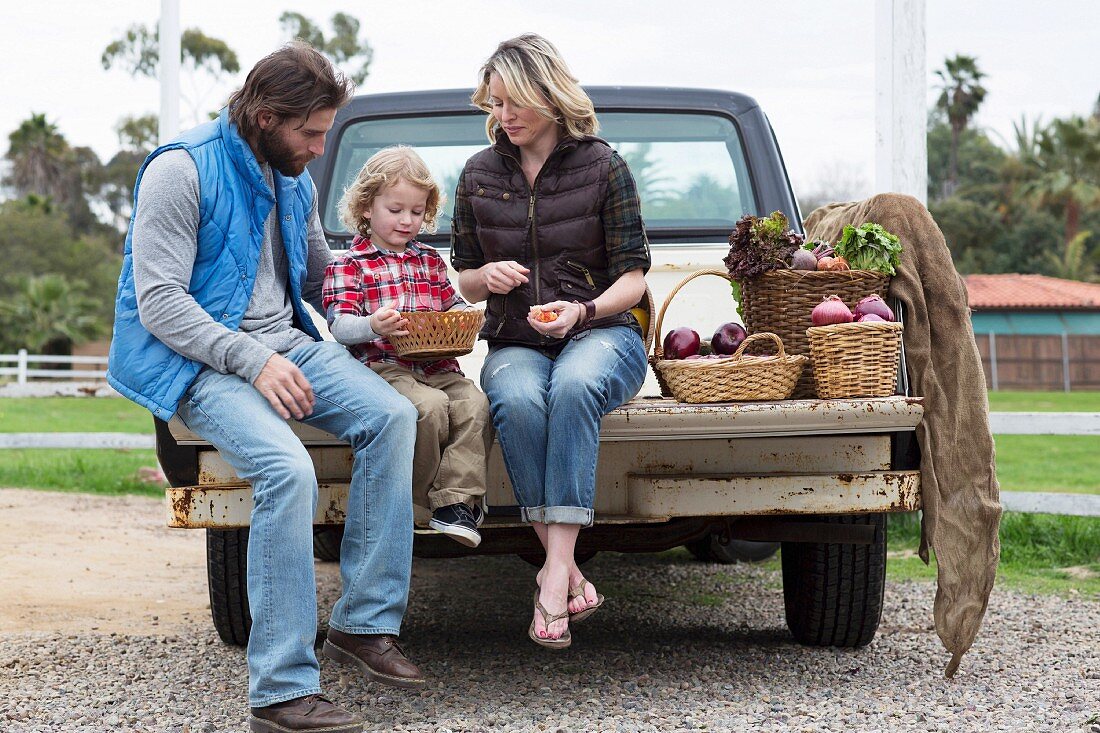Familie sitzt mit Obst & Gemüse in Körben auf Ladefläche eines Pickup