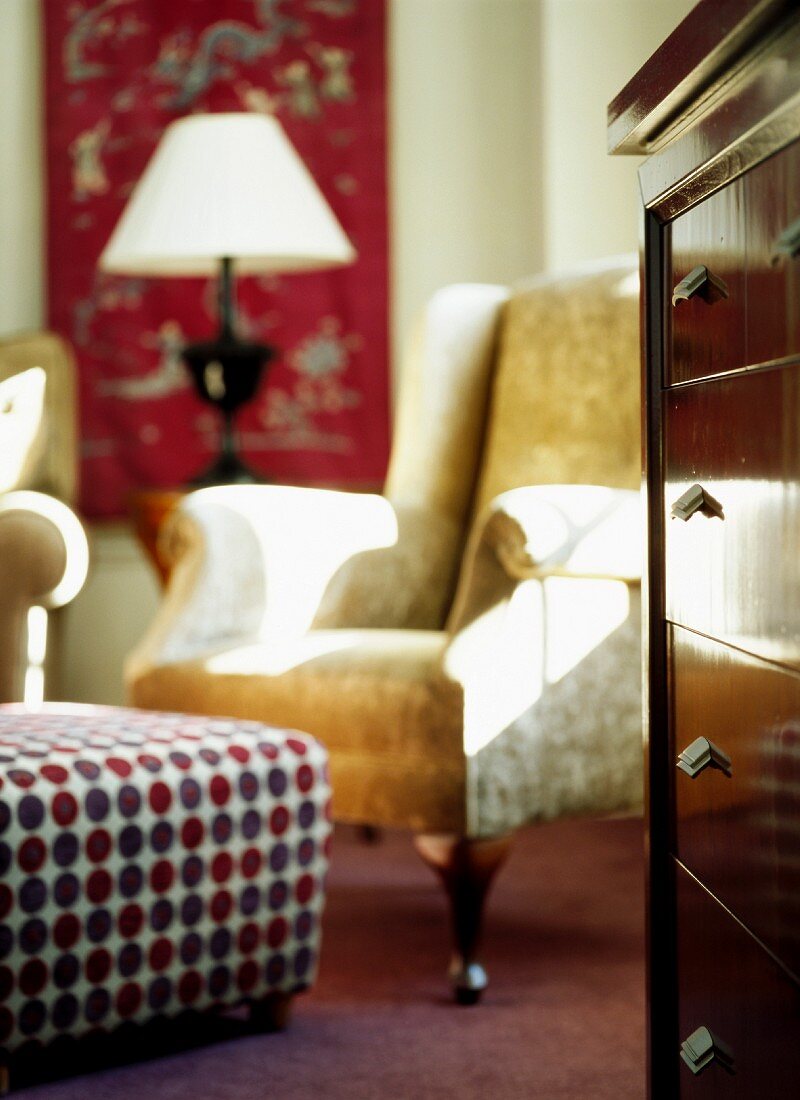 Patterned ottoman in front of antique leather armchair and chest of drawers in corner of living room