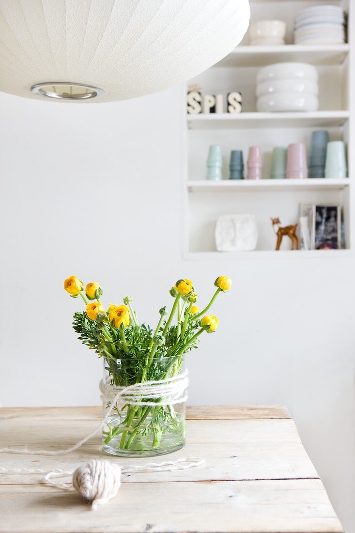 Blumenstrauss aus Ranunkeln in Glasgefäss mit Schnurdeko auf einem Holztisch, Ausschnitt einer Pendellauchte und im Hintergrund Regal in Wandnische