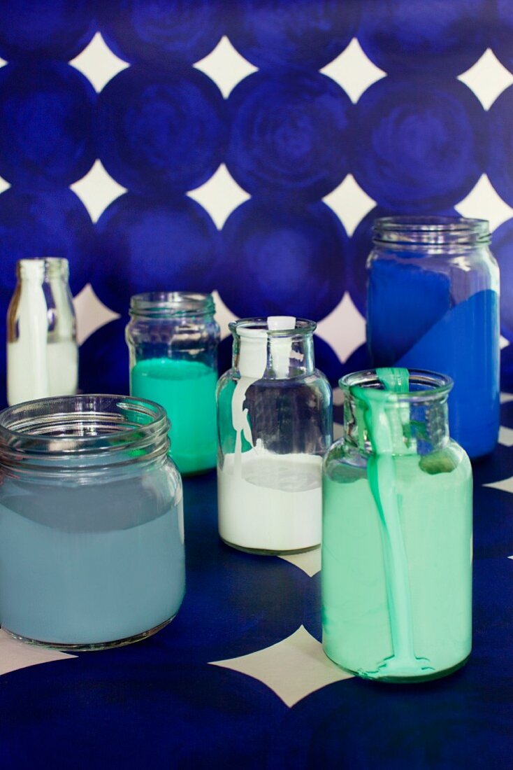 Various paints in glass jars on length of blue patterned wallpaper