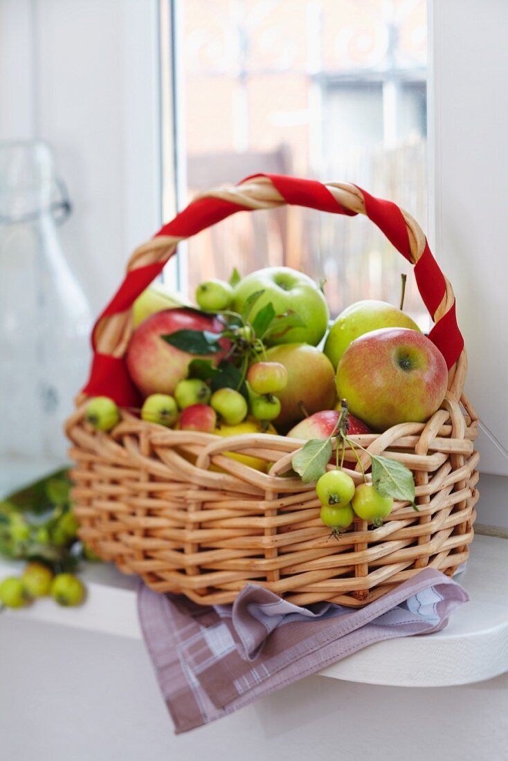 Korb mit Äpfeln und Zieräpfeln auf Fensterbank, Henkel mit rotem Band umwickelt