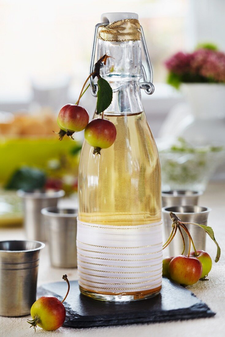 Apfellikör in dekorativer Bügelflasche mit Zieräpfeln, daneben kleine Zinnbecher