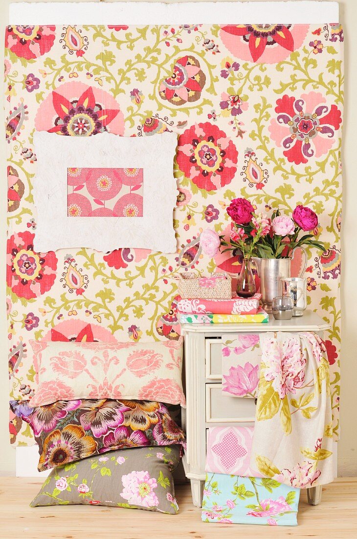 Stack of floral cushions below picture frame on pastel, patterned wall covering next to vintage cabinet draped with bolts of fabric