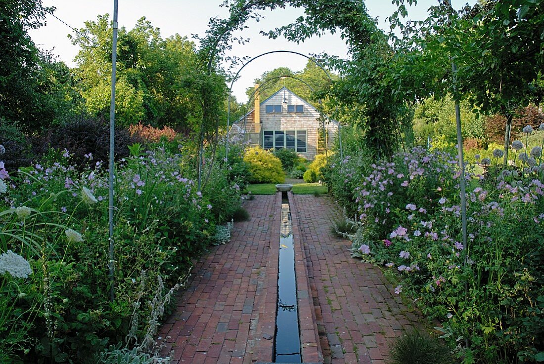 Ziegelsteinweg mit Wasserrinne gesäumt von lila Gartenblumen