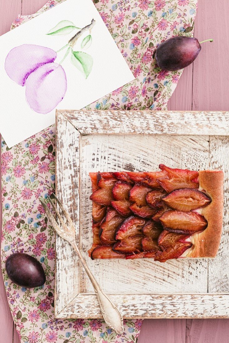 A slice of plum cake on a wooden tray and a picture of plums