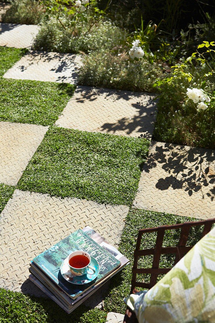 Chequered pattern of stone slabs and squares of lawn in sunny garden with flower bed to one side