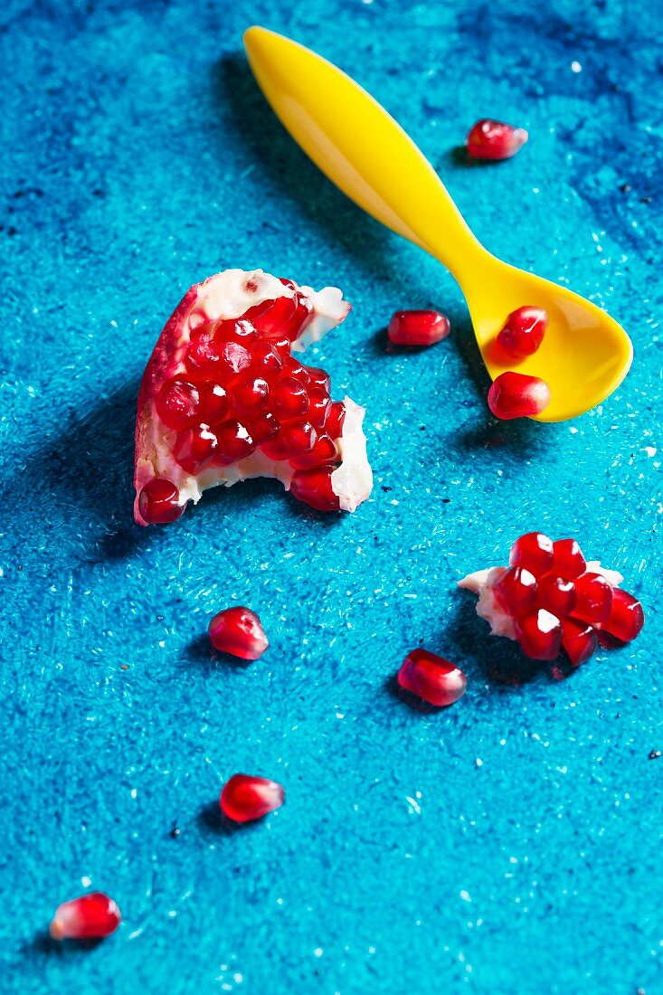 A chunk of pomegranate and pomegranate seeds on a turquoise surface with a yellow spoon