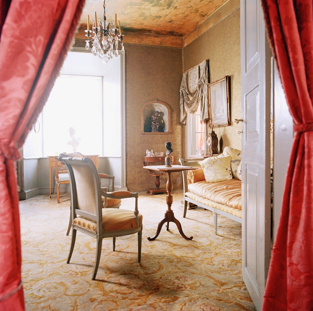 View through open door with curtains into elegant interior with carpet, antique seating & chandelier