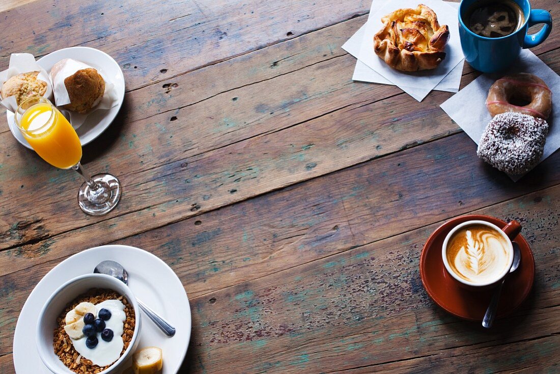 Frühstück auf Holztisch: Müsli mit Heidelbeeren und Joghurt, Orangensaft, Kaffee, Doughnuts und Muffins
