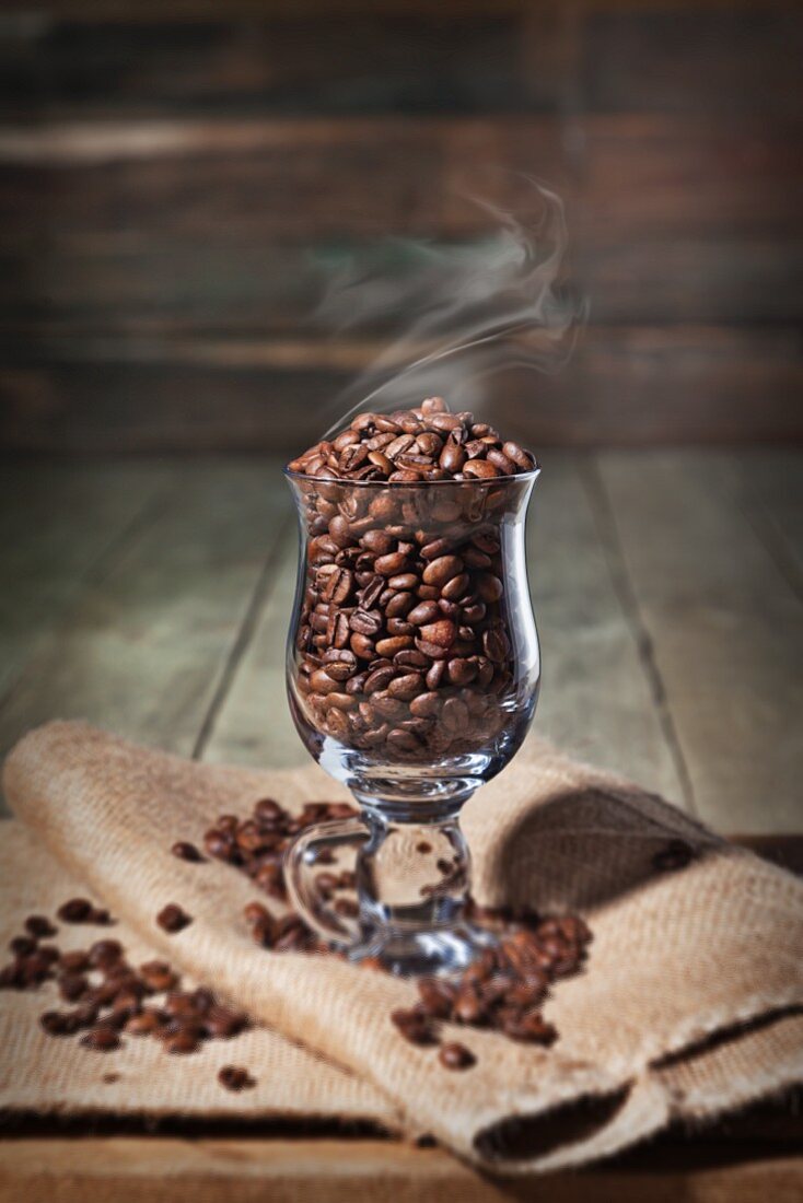 A Glass Coffee Cup with Steaming Coffee Beans on a folded Burlap Bag