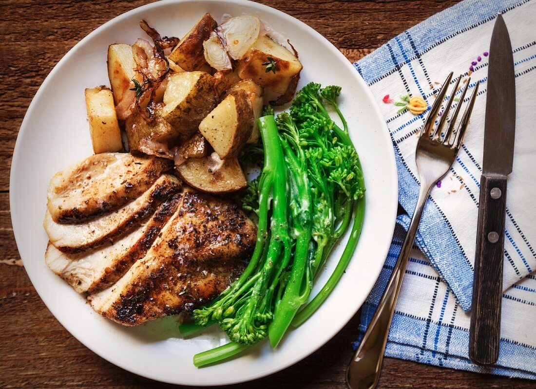 Sliced Grilled Chicken, Broccolini and Roasted Potatoes with Thyme on a White Plate set on a Wood Table with a Blue Plaid Napkin
