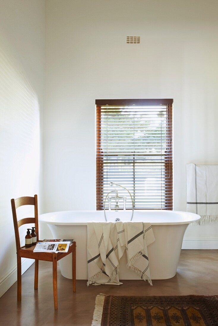 Freistehende Badewanne mit abgelegtem Handtuch vor Fenster und geschlossener Jalousie, an der Seite schlichter Küchenstuhl aus Holz mit Badutensilien