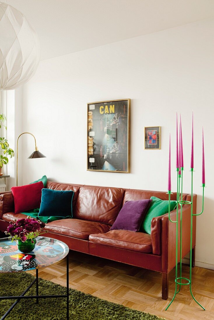 Beistelltisch mit buntem Tablett vor braunem Ledersofa, daneben grüner Metall Kerzenleuchter mit pinkfarbenem Kerzen in Wohnzimmerecke