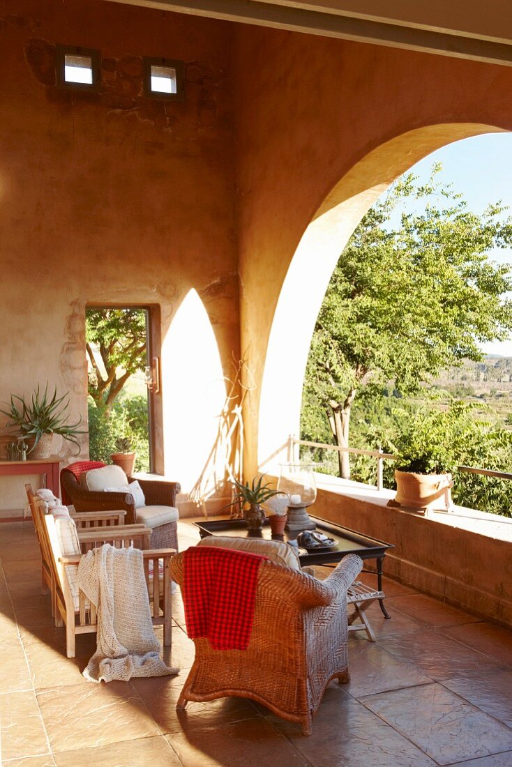 Loggia mit verschiedenen Sesseln und Couchtisch auf Steinfliesenboden und Blick durch Rundbogen in sonnigen Garten