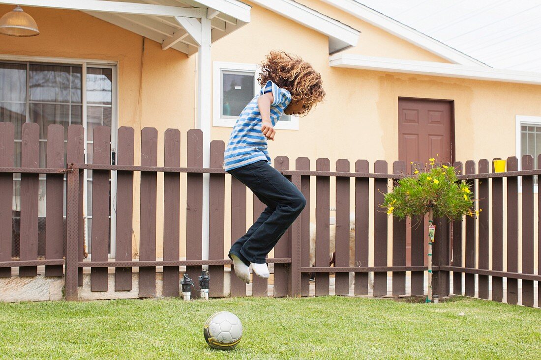Kind beim Ball spielen im Garten mit Holzzaun vor apricot getöntem Wohnhaus