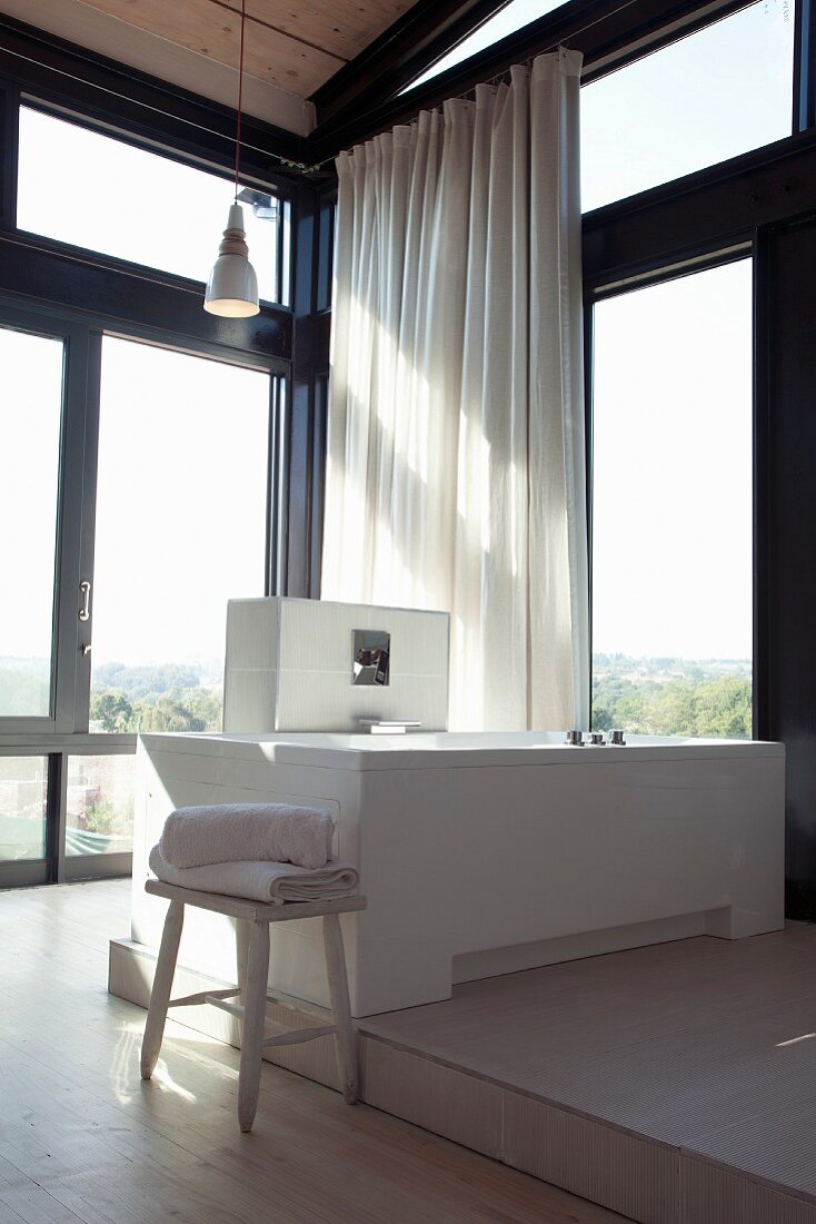 Bathtub on platform in corner of room with glass walls in contemporary house with white curtain in front of window