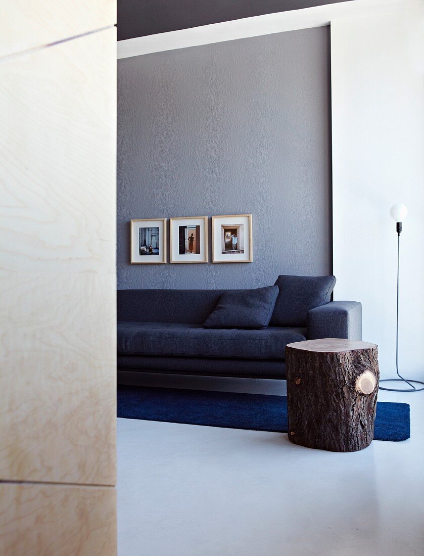 Elegante, anthrazitfarbene Couch an grauer Wand, darunter ein dunkelblauer Teppichläufer; ein rustikaler Holzstamm dient als Beistelltisch