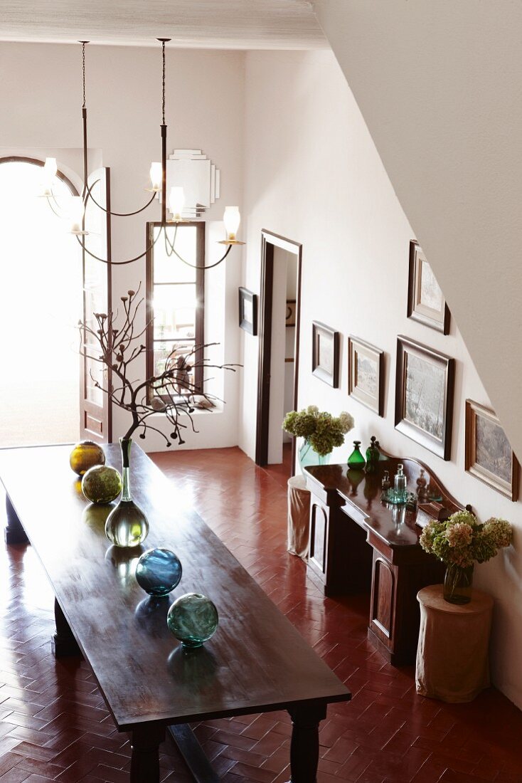 Row of glass spheres on long wooden table, vases of dried flowers and collection of pictures on wall of large foyer