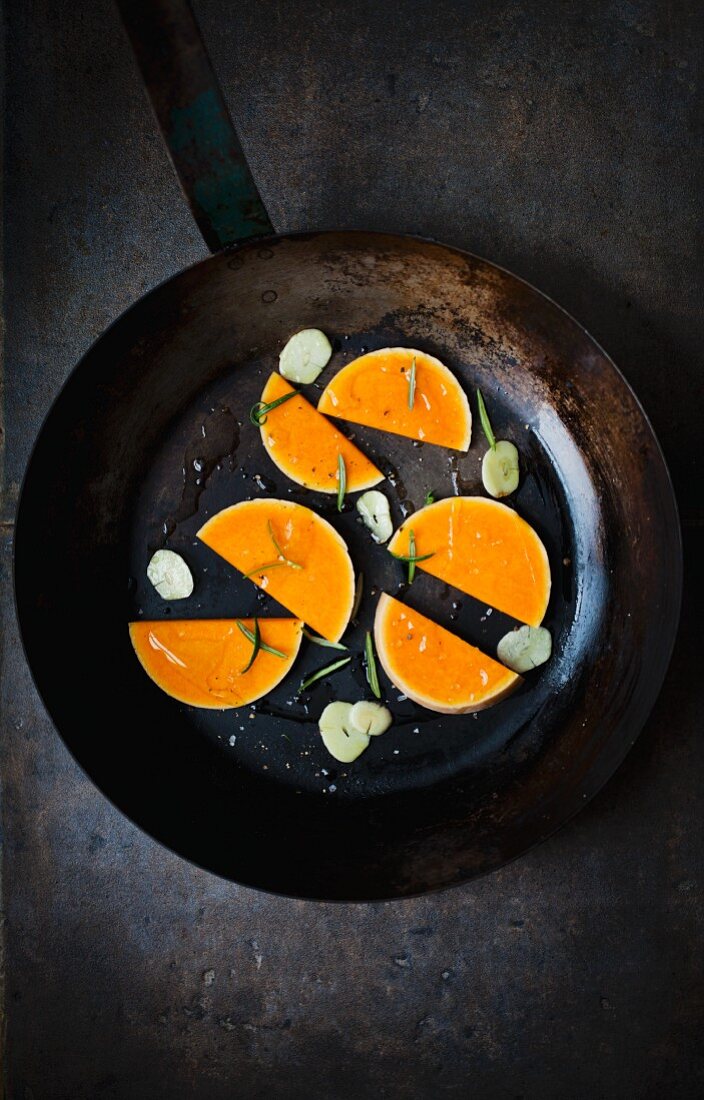 Slices of raw butternut squash with garlic and rosemary in an old frying pan