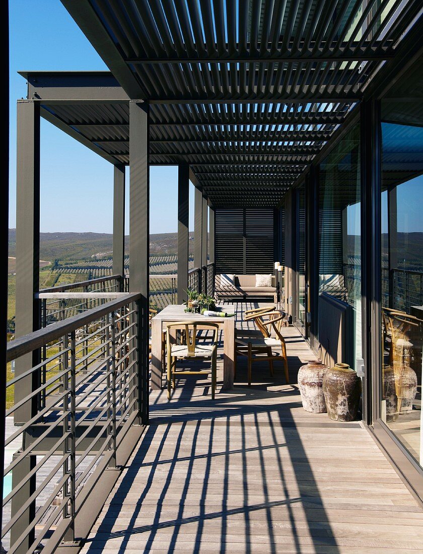 Sunny balcony with metal supports, metal balustrade and wonderful panoramic view; simple wooden table, chairs and African floor vases